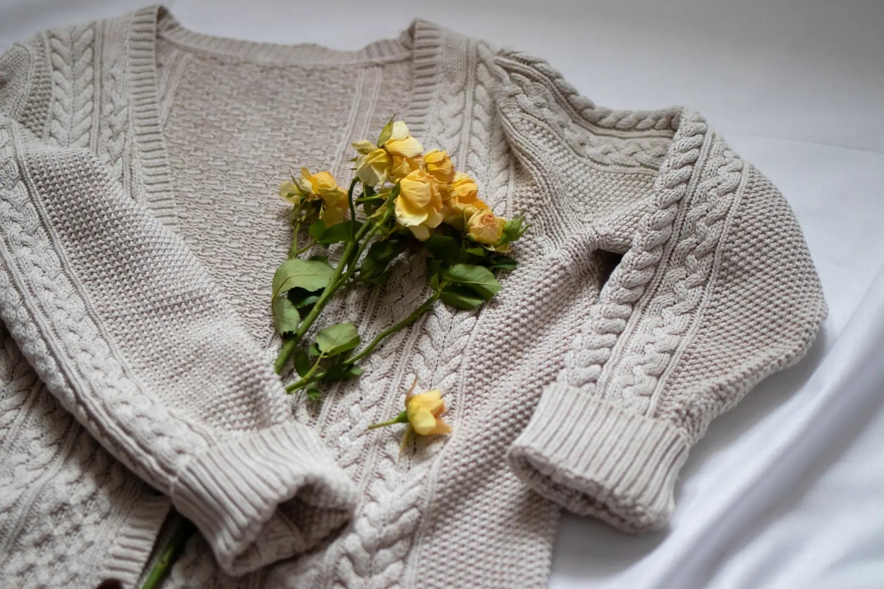 A cream cable-knit sweater laid flat with a few small yellow roses on it